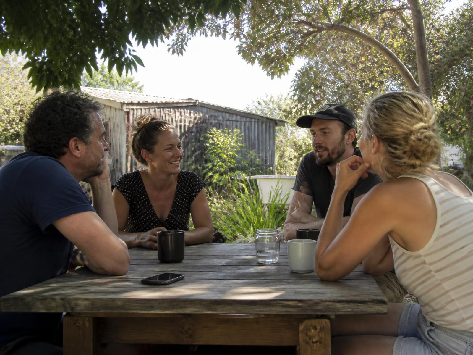 Four people talking around a wooden table outdoors under dappled tree shade.